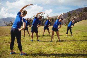 A group of people wearing blue t shirts and black shorts or leggings. They are exercising in a grass field with a wooded hillside in the distant background behind them.