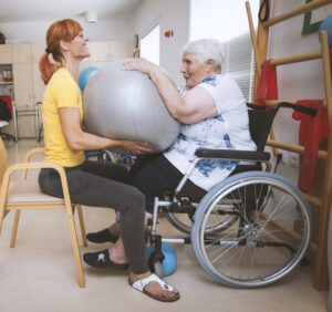 A female fitness instructor sitting on a chair and wearing a yellow t-shirt and black leggings. Opposite her is an older female wheelchair user wearing a white top and black leggings. The fitness instructor is holding a grey exercise ball from below, and the older woman has her hands on top of the ball pushing downwards.