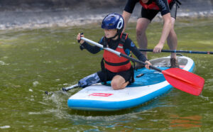 A boy with a prosthetic leg sitting on a paddleboard and wearing a blue helmet and red lifejacket. He is holding a red paddle.